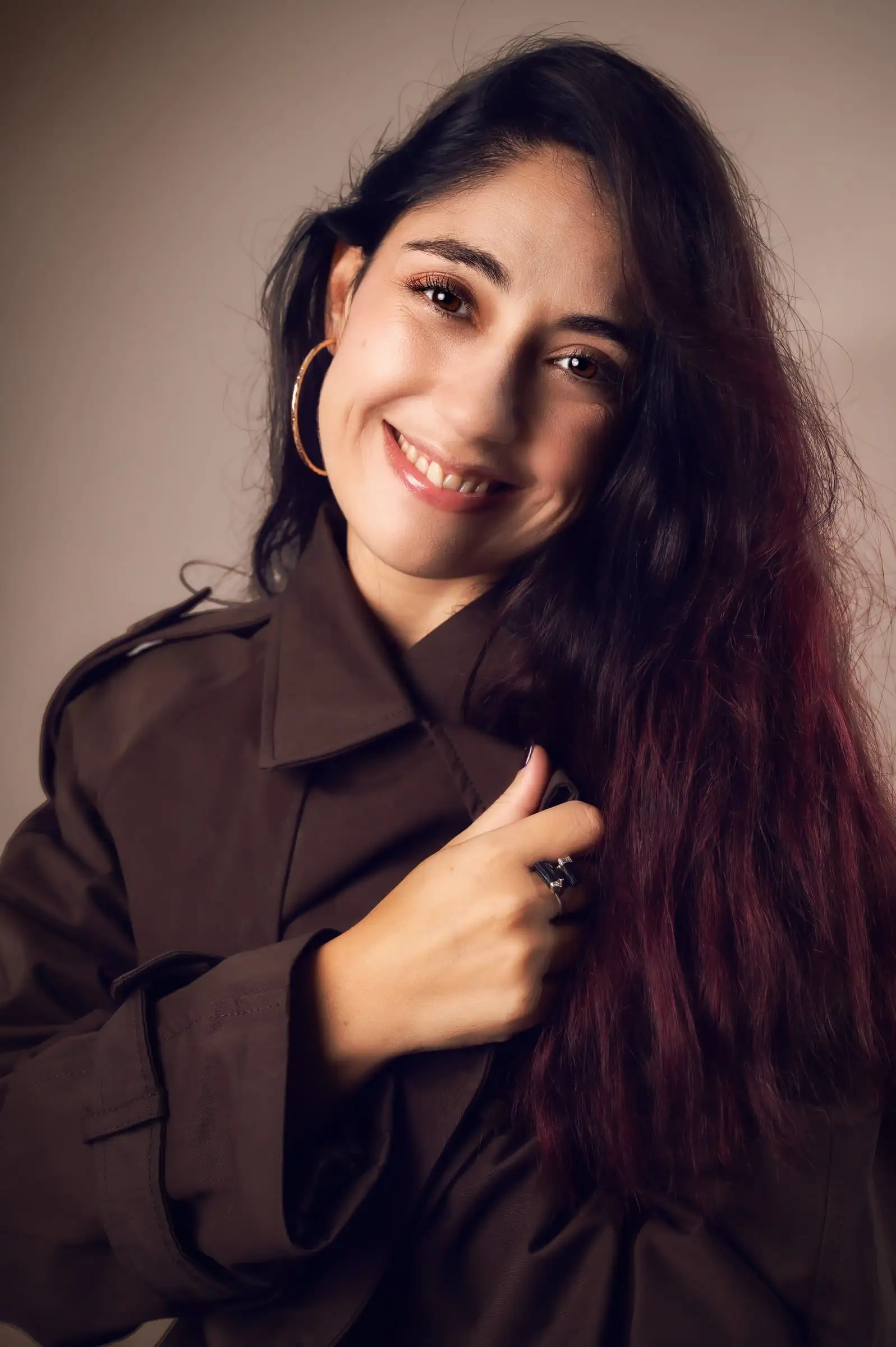 Mujer sonriente con camisa oscura y una mano junto al cabello, sobre fondo cálido.